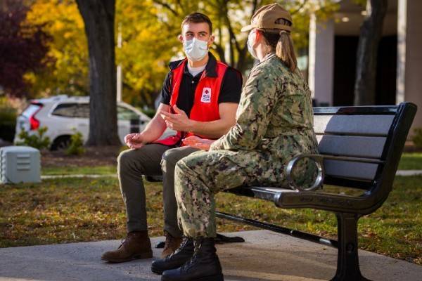 Red Cross volunteer and military service member, wearing masks, sitting on park bench and talking to each other.