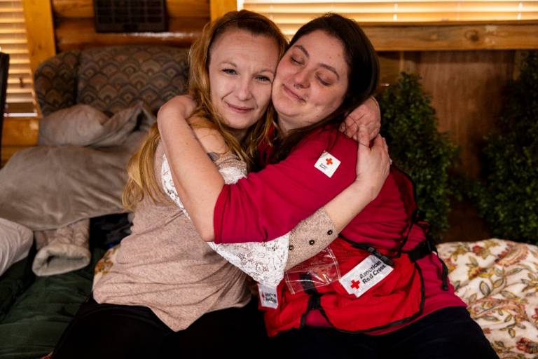 woman in tan shirt hugs woman in Red Cross vest in disaster shelter
