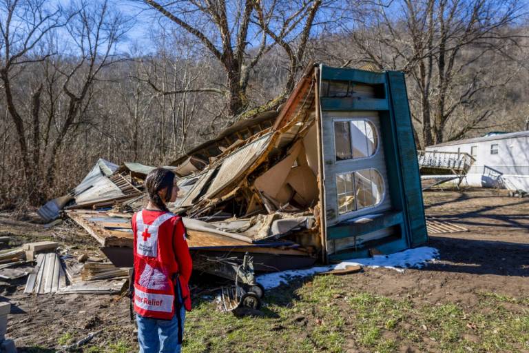 woman in Red Cross vest and jeans looks out over a destroyed mobile home