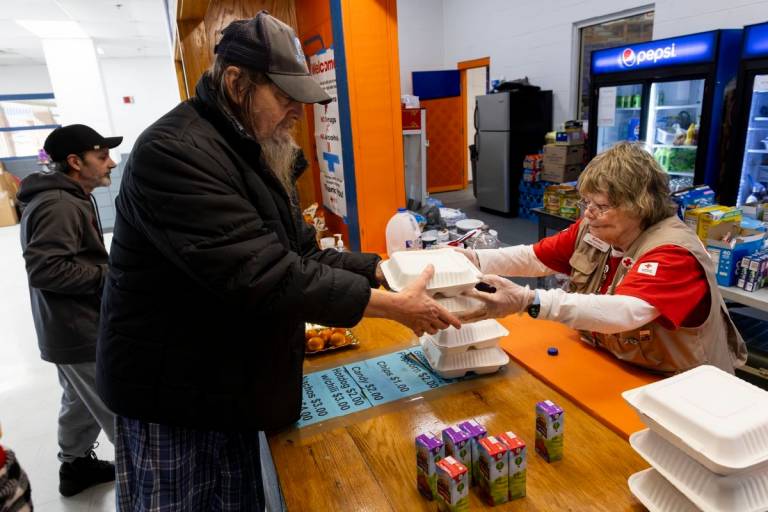 two men in dark jackets in front of a table get a warm meal from woman in Red Cross vest serving food from behind the table