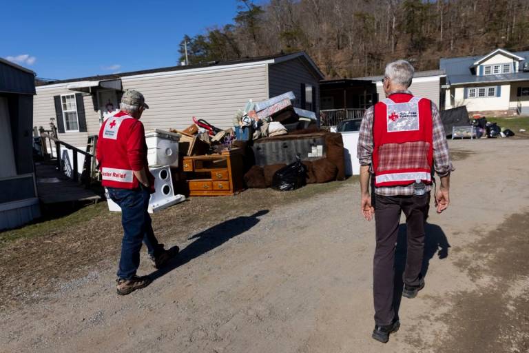 two men in dark pants and Red Cross vests walk through damaged neighborhood