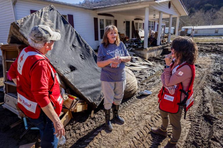 a man and woman in Red Cross vests with woman in rain boots and old clothes talk in front of piles of items removed from the damaged home in the background