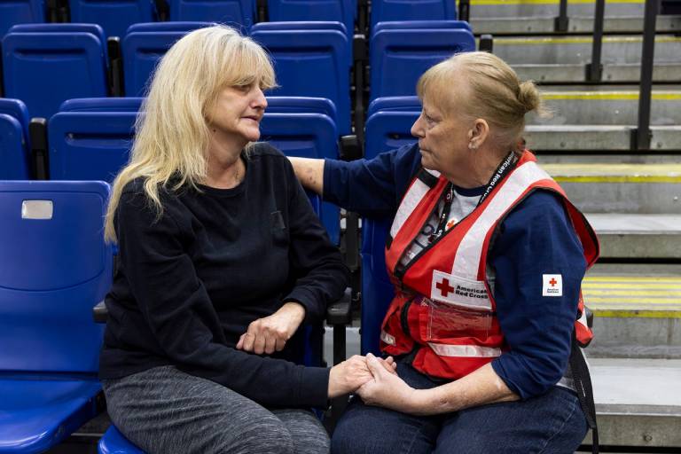 Two women sit and talk in a disaster shelter, one in black shirt and one in Red Cross vest