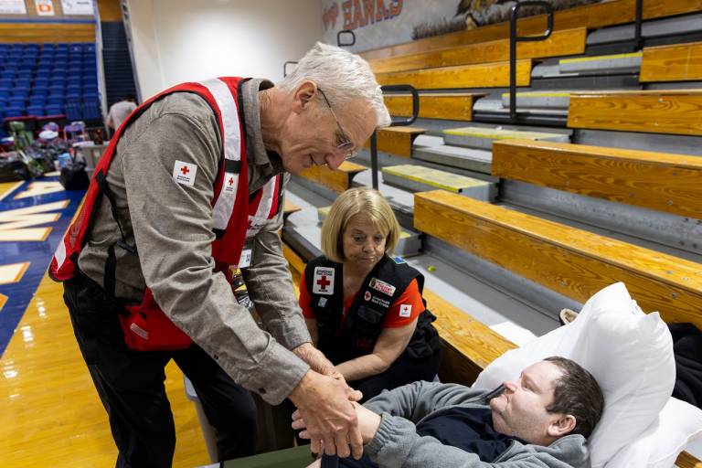 A man and woman check on another man resting on a cot in a Red Cross disaster shelter