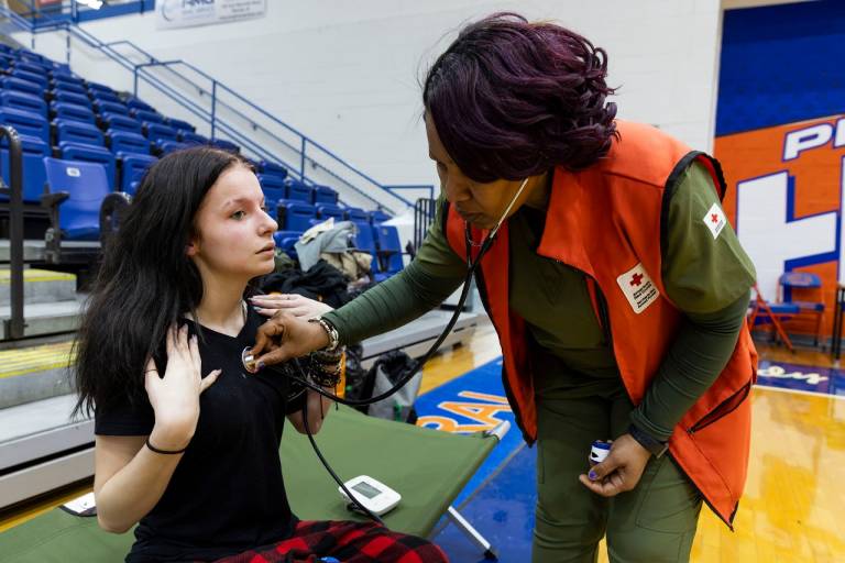 A woman in a Red Cross vest with a stethoscope checks on a young woman in a disaster shelter