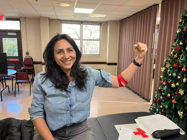 young woman sits on blood donor bed dressed casually with her right arm raised into the air