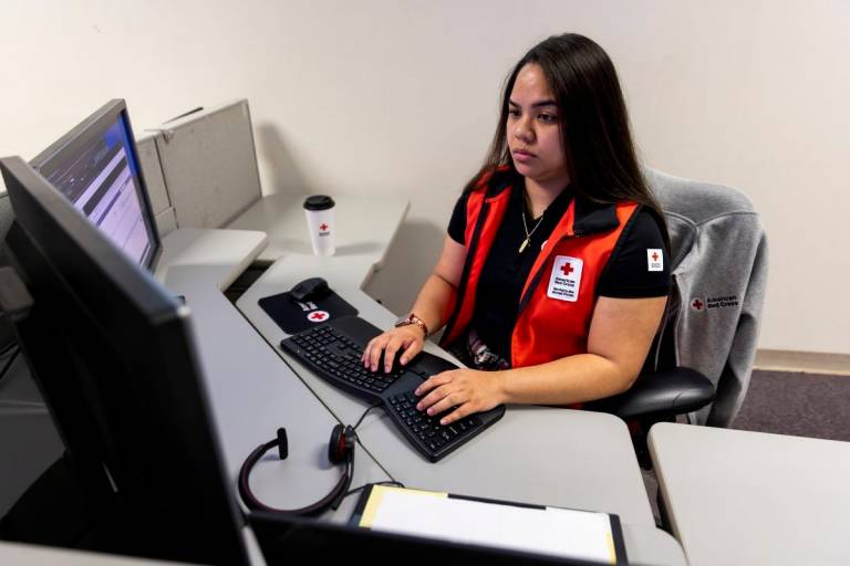 young woman in Red Cross vest sits at a computer desk