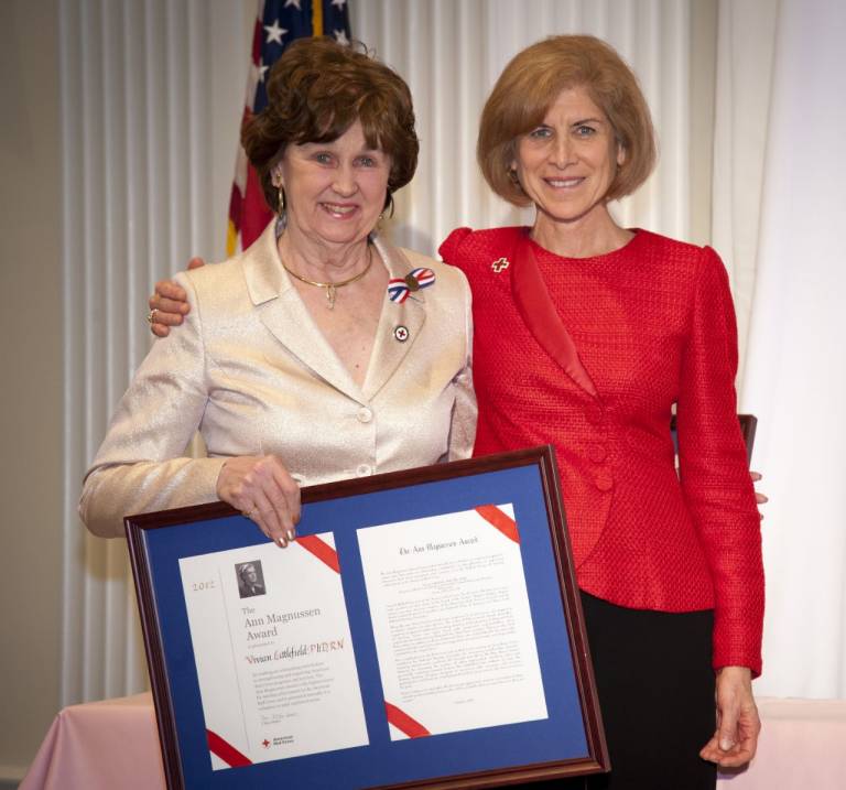 two women, one in white suit holding award, the other in red jacket