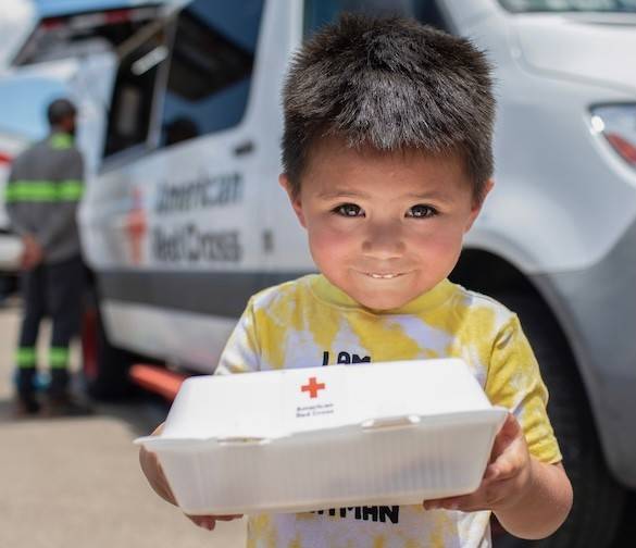 A small child standing in front of an American Red Cross emergency response vehicle smiles while holding up a container of food. There is a logo in the top right corner reading Giving Day - American Red Cross, March 26