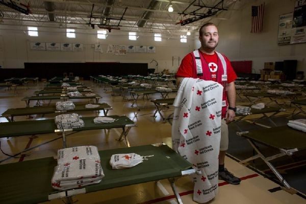 Red Cross volunteer holding a Red Cross blanket and standing in a shelter surrounded by shelter cots.