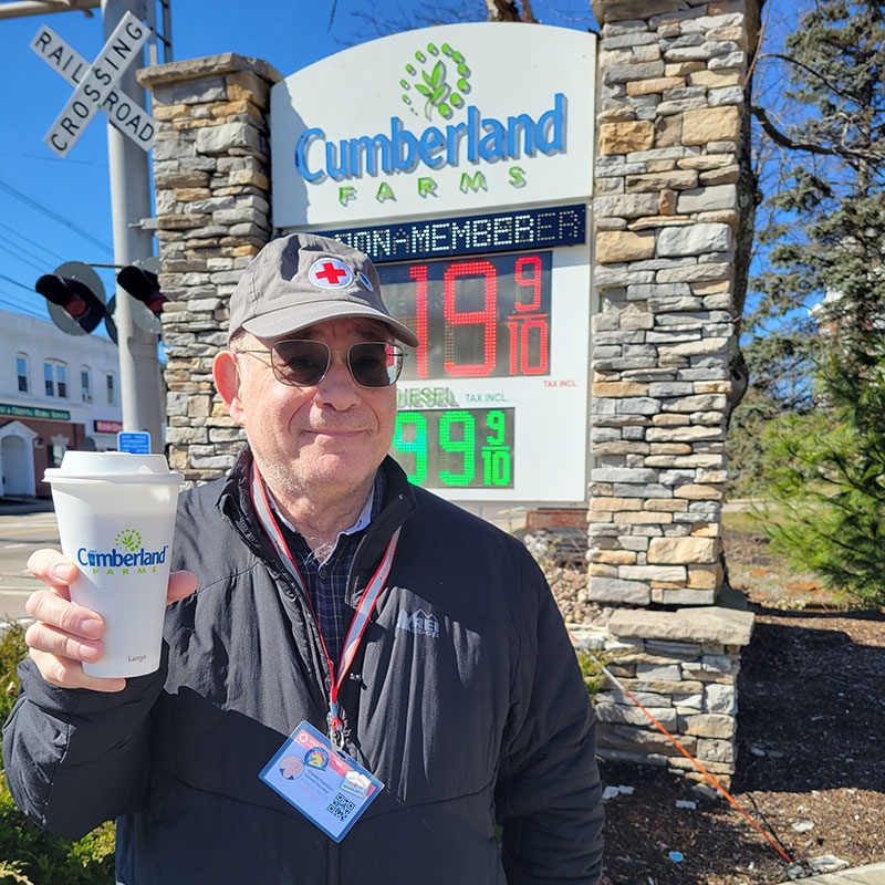 Red Cross volunteer holding up a cup of coffee in front of the Cumberland Farms sign by the street.