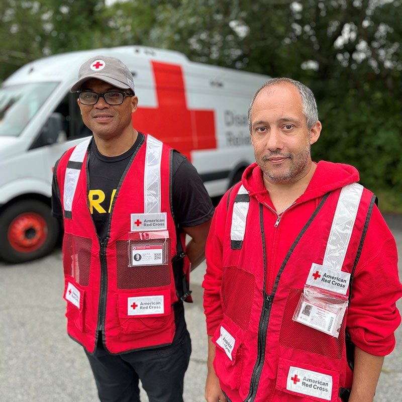 Two Red Cross volunteers in Red Cross vests and a Red Cross van behind them, smile for picture.
