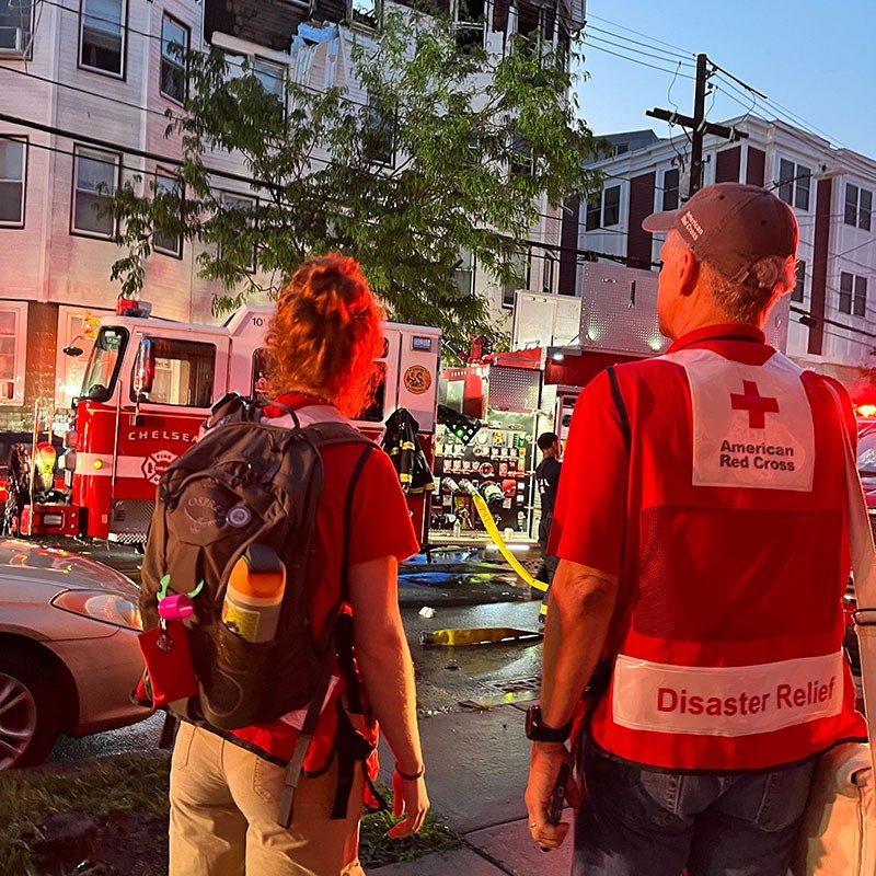 Red Cross volunteers looking at fire engine next to a four story building with fire damage on top floor.