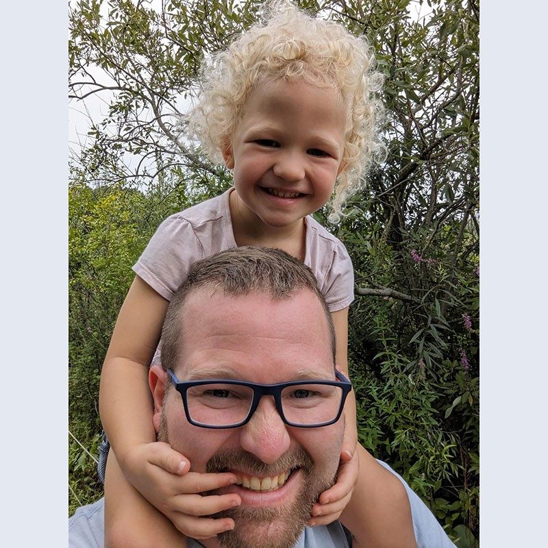 Child sitting on dad's shoulders and smiling for a selfie.