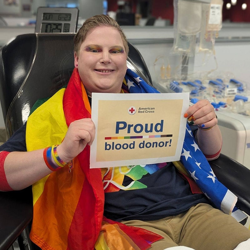 Kaleb sitting in chair with pride flag around shoulders and holding a sign that says, Proud Blood Donor!