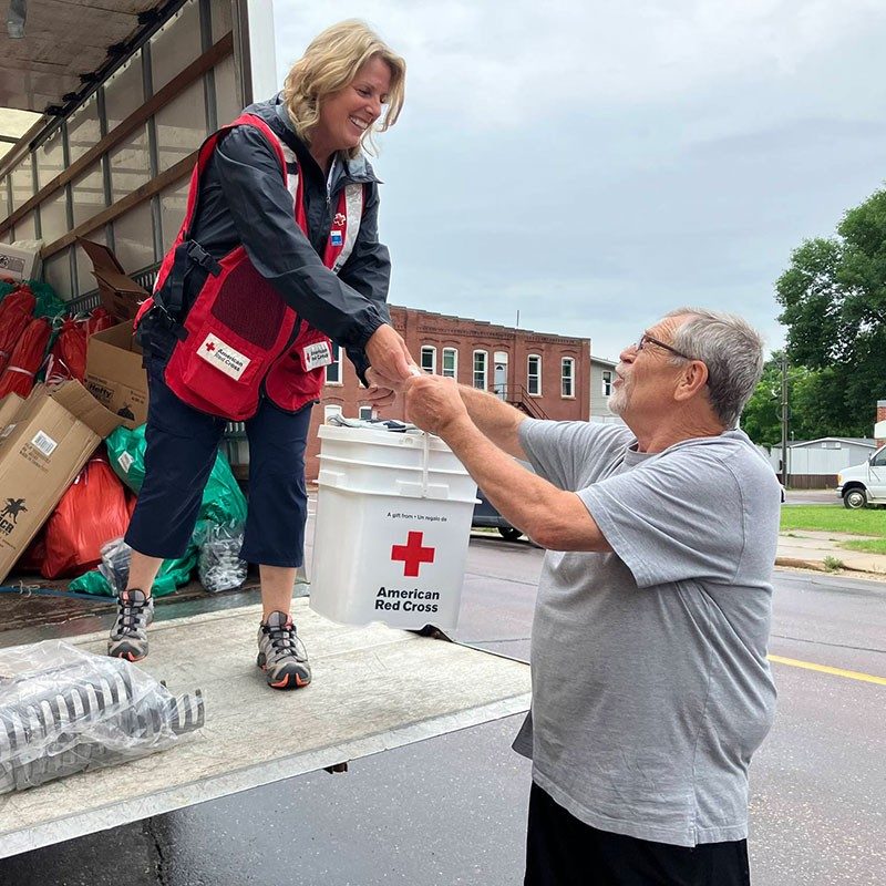 Red Cross volunteer standing on lift gate of moving truck and handing a 5 gallon Red Cross bucket to a person below.