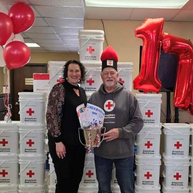 Bill “Waz” Waerzeggers, holding a pail with paper that says congratulations, standing next to a Red Cross employee in a room with baloons and stacks of American Red Cross 5 gallon buckets behind them.