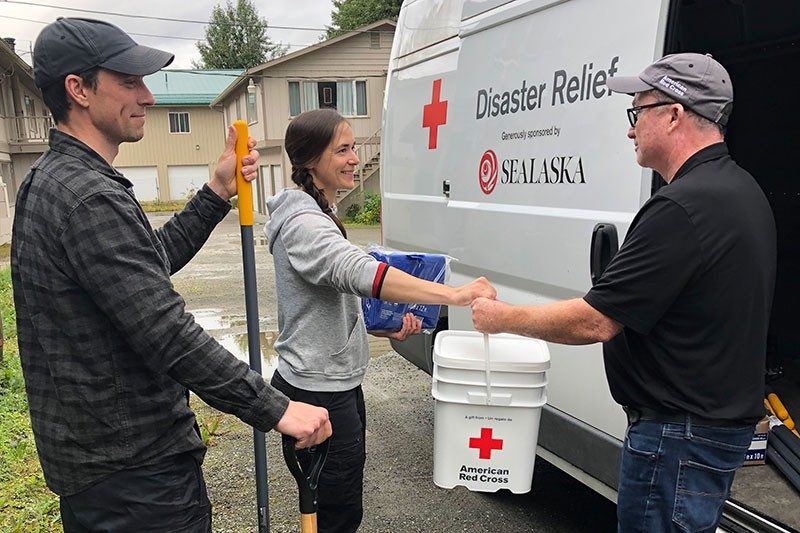 American Red Cross volunteer handing an American Red Cross 5 gallon bucket to a person who's standing next to another person who's holding a rake and shovel.