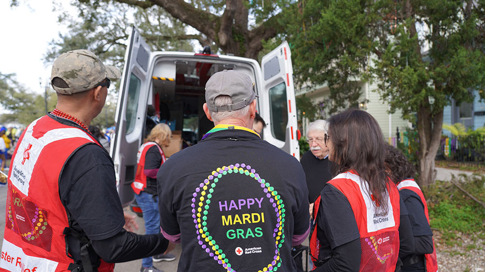 Group of Red Cross volunteers wearing Red Cross vests and happy mardi gras shirt, gathered around the back of a Red Cross van.