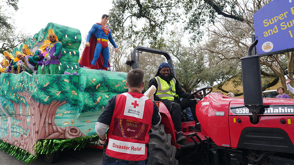 Red Cross volunteer wearing a Red Cross vest handing food to person on tractor that's pulling a mardi gras float.