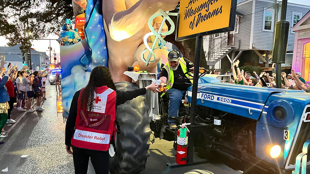 Red Cross volunteer wearing a Red Cross vest handing food and drink to person on tractor that's pulling a mardi gras float.