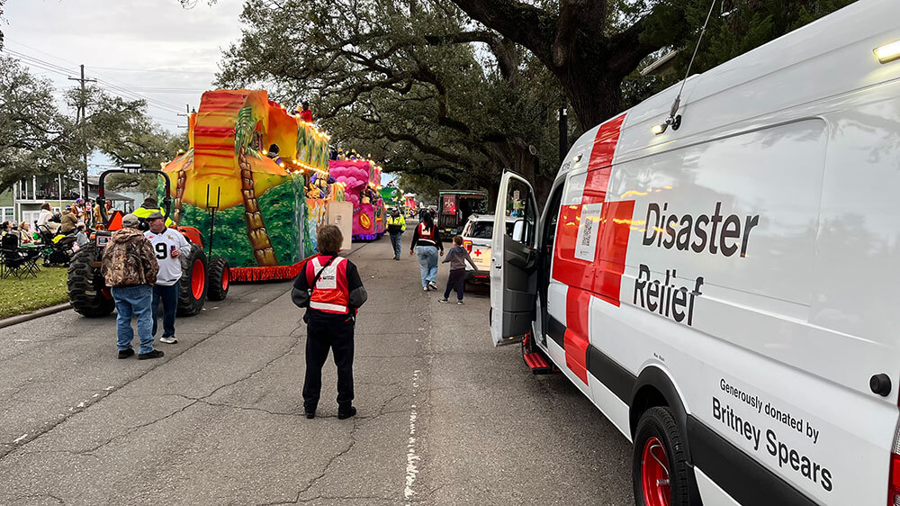 Red Cross volunteer standing in street standing next to a Red Cross van with mardi gras floats lined up.