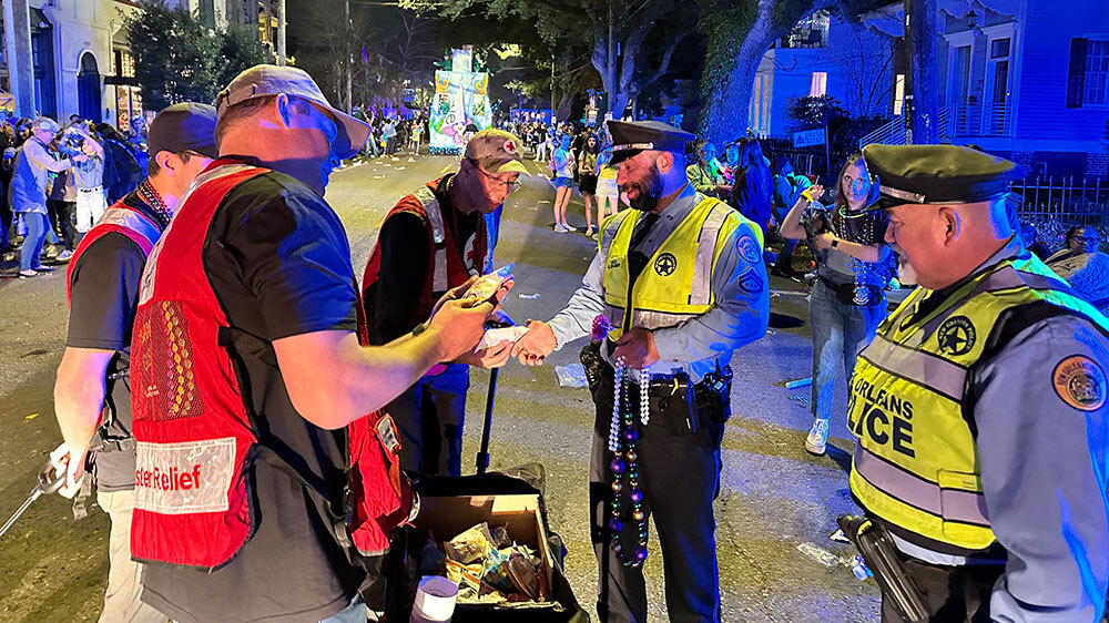 Red Cross volunteers delivering snacks to police officers at mardi gras parade with crowd of people alomg the street.