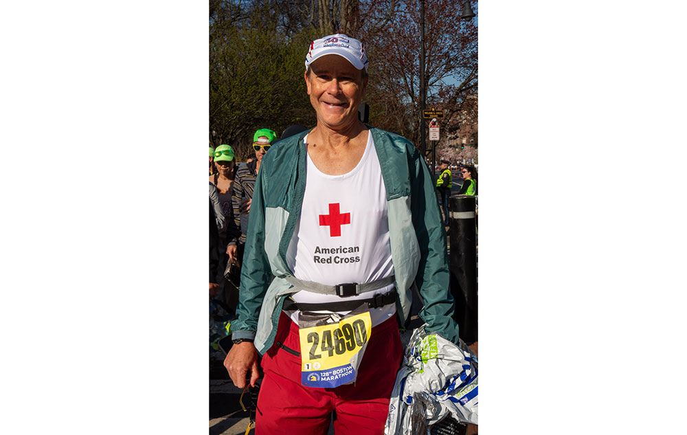 Marathon runner smiling for camera wearing hat, jacket, Red Cross shirt and marathon number on waist.