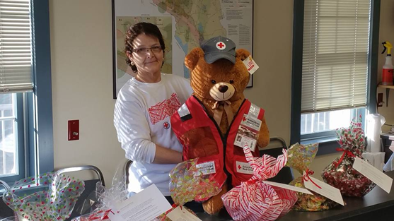 Red Cross volunteer holding large stuffed bear that's wearing a Red Cross hat and vest.