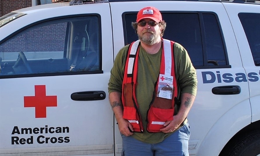 Keith McAuliffe wearing Red Cross hat and vest, standing next to a Red Cross vehicle.