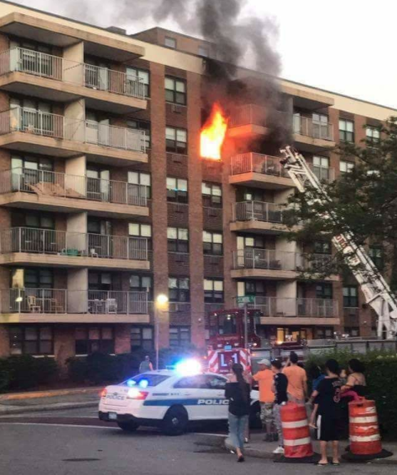 Apartment building fire on the 5th floor with fire engine, police car and people standing outside.