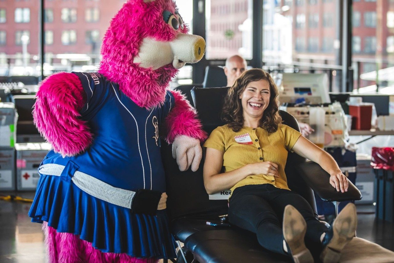 Person sitting in chair ready to donate blood with a mascot standing next to chair.