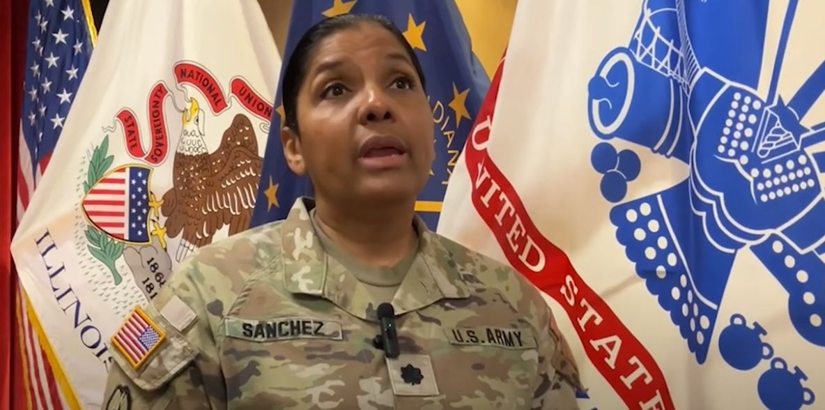 Military member in camo uniform standing in front of multiple flags.