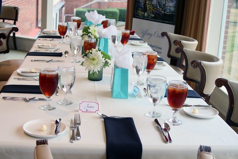 Long rectangle dinner table set with filled glasses, plates, silverware, flowers and gift bags.