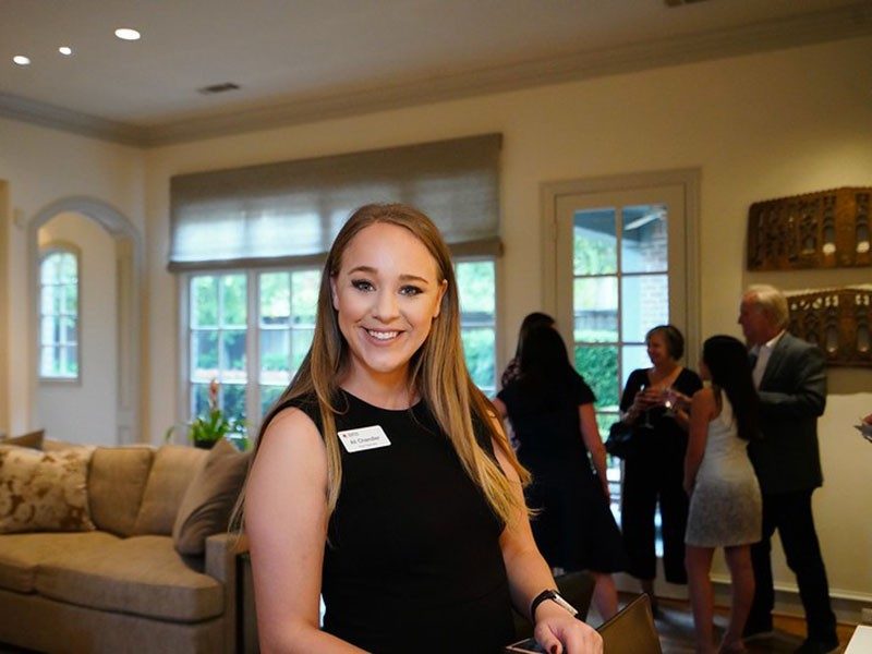 Person inside house smiling with group of people talking to each other in the background.