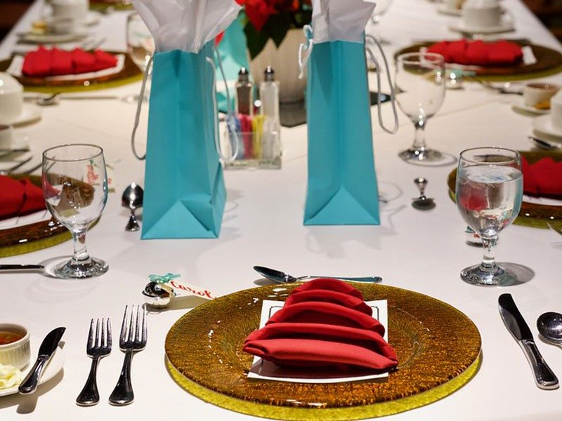 Dinner table set with filled glasses, plates, silverware, flowers and gift bags.