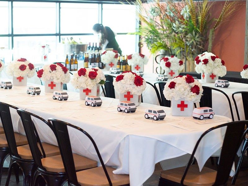 Long rectangle dinner table with multiple small American Red Cross van replicas and red and white flowers in vases with American Red Cross logos on them.