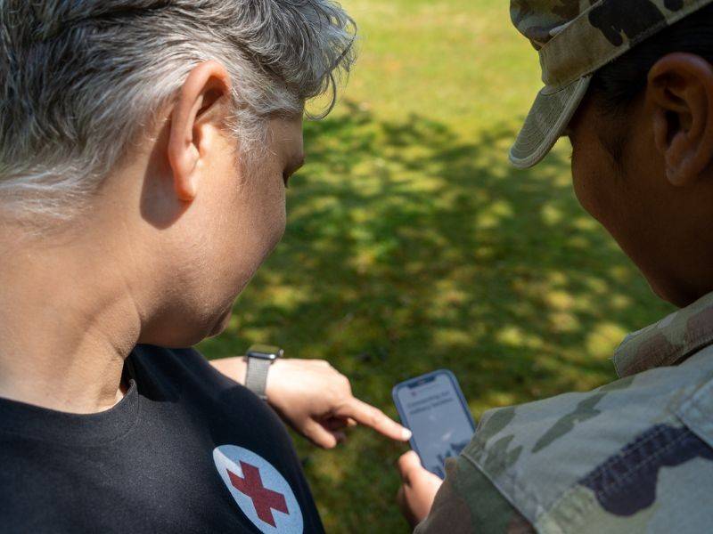 Military service member holding and looking at a smartphone with Red Cross volunteer pointing at phone.