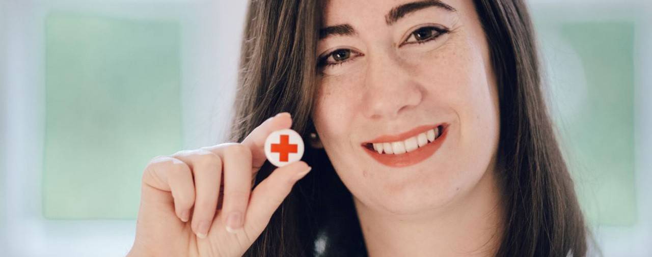 smiling woman with brown hair holds her Red Cross pin to the camera