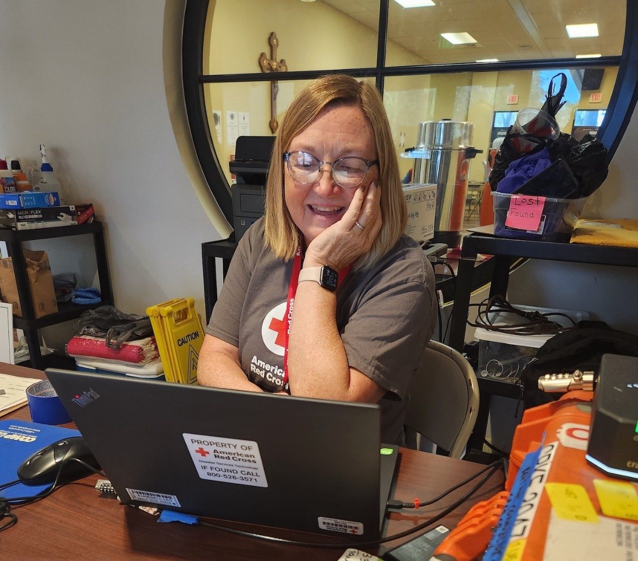 Suzanne Garrett sits at a cluttered brown desk working on a black laptop with an American Red Cross sticker while on deployment.