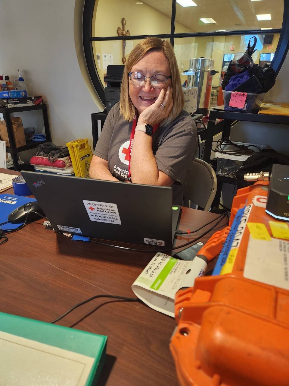 Suzanne Garrett sits at a cluttered wooden desk working on an American Red Cross branded laptop during a deployment.