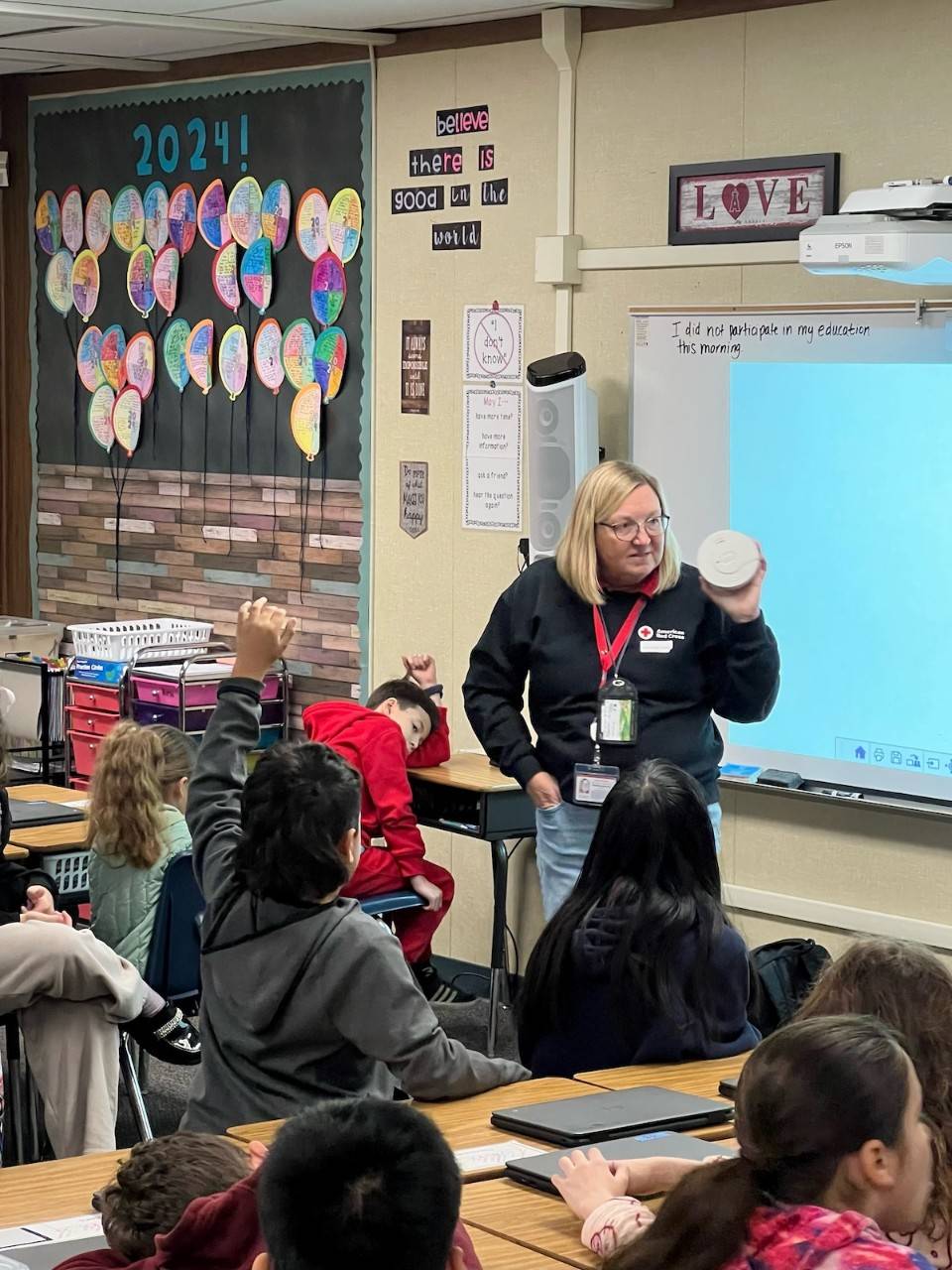 Suzanne Garrett holds a smoke detector up as she stands in front of a classroom full of young children sitting at their desks. 