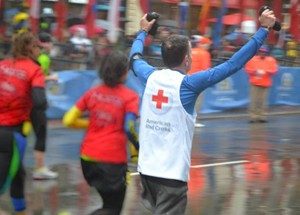 marathon runner wearing red cross shirt holding hands up after crossing finish line.