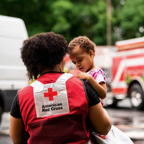 An American Red Cross Disaster Relief volunteer holds a small child while they look out over a fire truck on the road.