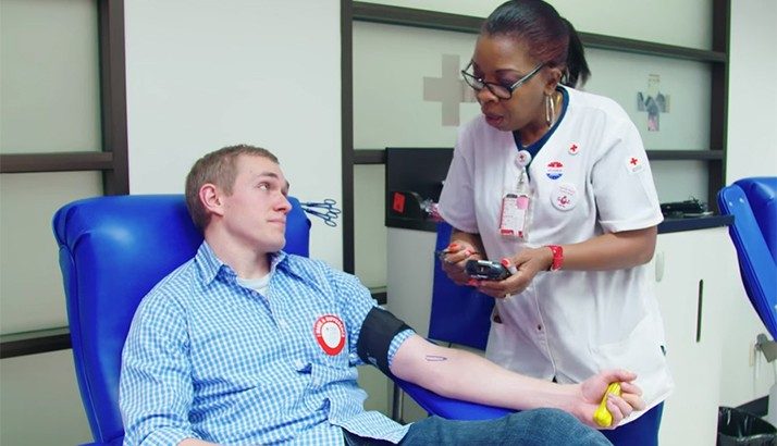 Man sitting in a chair giving blood and speaking with a phlebotomist.